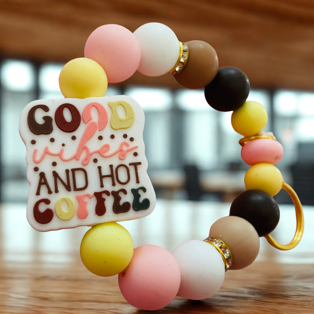 Colorful beaded bracelet with a square charm reading 'Good vibes and hot coffee' on a blurred indoor background.